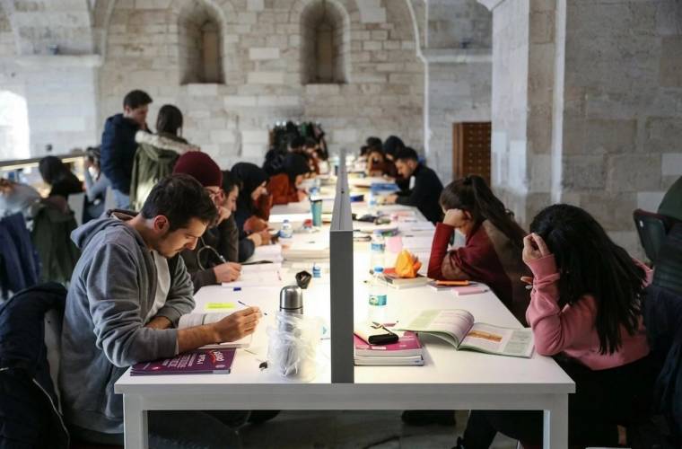Taş duvarlı salonda, uzun masalarda oturan gençler kitap ve defterlerle çalışıyor.