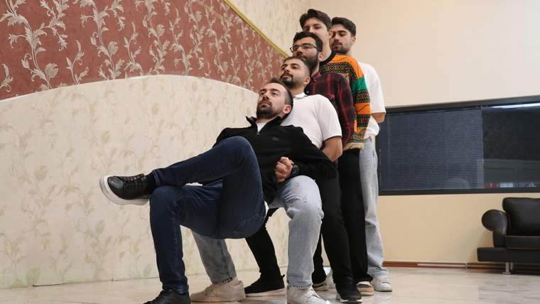 Bir salonda beş kişi, birbirlerinin arkasında sıralanarak bacaklarını öne uzatıp duruyor.