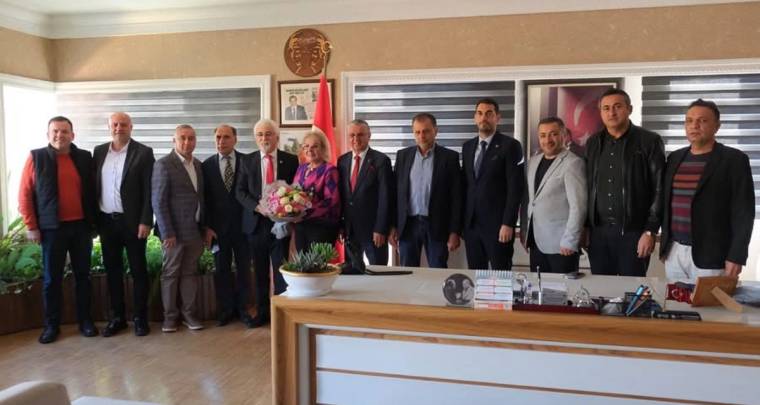 Bir ofiste masa önünde yan yana duran kalabalık grup; ortadaki kadın elinde çiçek buketi tutuyor.