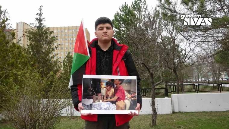 Kırmızı montlu genç bir kişi, Filistin bayrağı ve üzerinde fotoğraf bulunan bir pankart tutuyor.