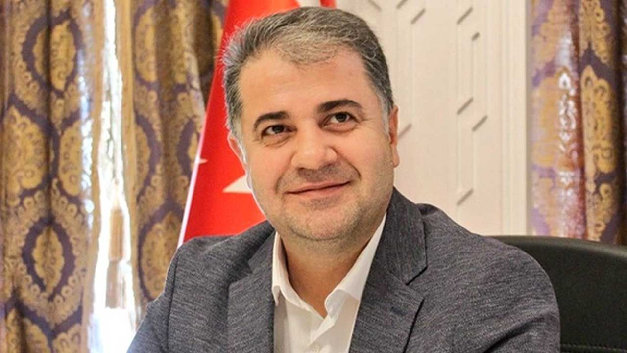 Gri ceketli bir kişi, arkasında Türk bayrağı ve desenli perdeler bulunan bir ortamda oturuyor.