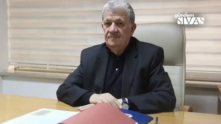 Gri saçlı ve bıyıklı bir kişi, ofis ortamında masanın arkasında oturuyor; önünde dosyalar bulunuyor.