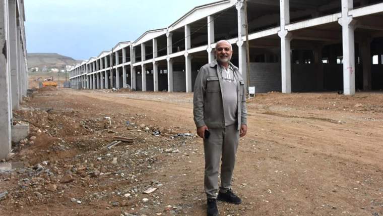 İnşaat halindeki uzun bir yapı önünde, gri kıyafetli bir kişi toprak zeminde ayakta duruyor.