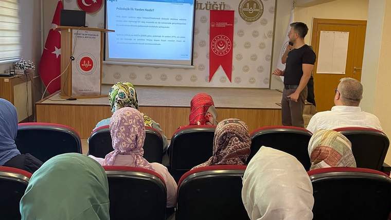 Toplantı salonunda bir kişi sunum yaparken, başörtülü ve başı açık katılımcılar ekrana bakıyor.