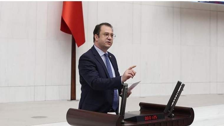 Kürsüde konuşan bir kişi, elindeki kâğıdı tutarak işaret ediyor; arkasında Türk bayrağı görülüyor.