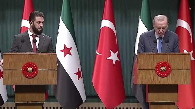 Yan yana kürsülerde duran iki erkek konuşmacı, aralarında Türkiye ve Suriye bayraklarıyla görülüyor.