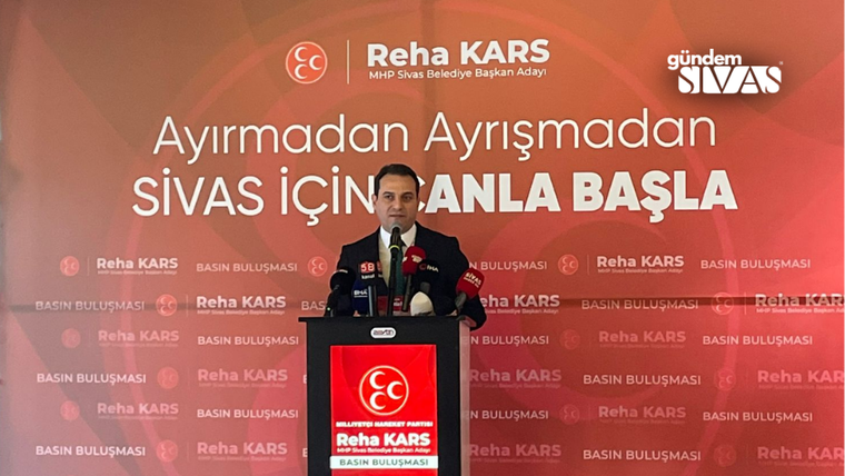 Kırmızı kürsüde konuşan bir kişi, arkasında “Reha Kars” ve “Sivas için planla başla” yazılı fonda duruyor.