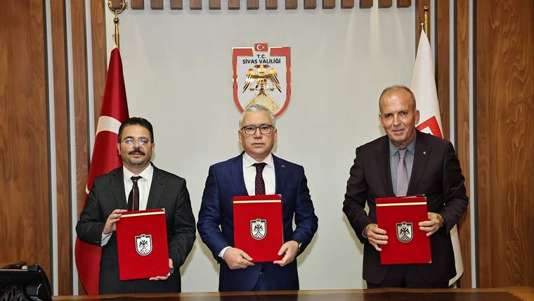 Üç kişi, masanın üzerinde bulunan dosyalarla birlikte poz veriyor. Arkada Türk bayrağı ve Sivas Valiliği arması var.