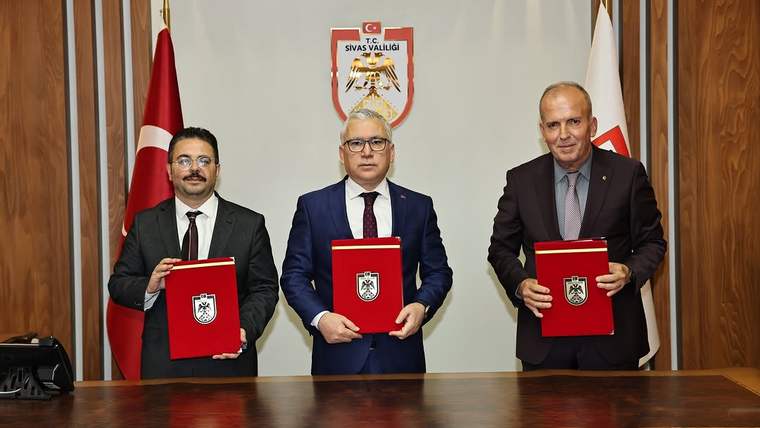 Üç kişi, masanın üzerinde bulunan dosyalarla birlikte poz veriyor. Arkada Türk bayrağı ve Sivas Valiliği arması var.