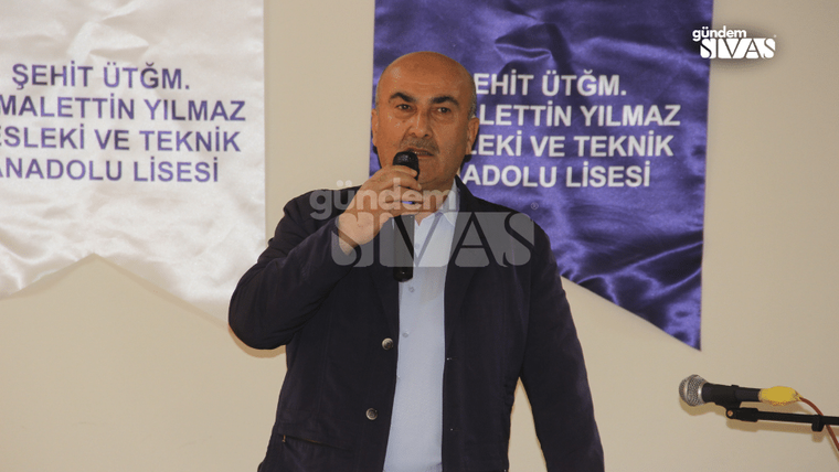 Bir kişi elindeki mikrofonla konuşuyor; arkasında lise adının yazılı olduğu afiş, yanında ikinci mikrofon görülüyor.
