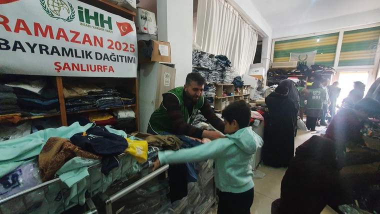 Bir çocuk, yardım dağıtımında bir yetişkinle birlikte giysi alıyor. Arka planda kalabalık bir ortam var.