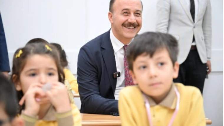 Sınıfta iki öğrencinin arkasında oturan bıyıklı bir adam gülümserken, çocuklar sıralarında görülüyor.