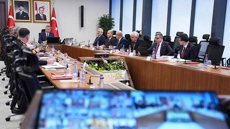 Bir toplantı odasında, masanın etrafında oturan kişiler ve ekranda video konferans görüntüleri yer alıyor.