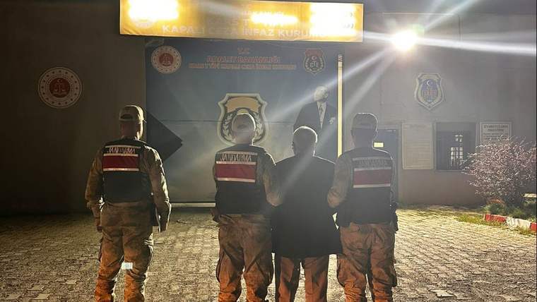 Gece, bir kamu binası önünde üç üniformalı görevli ve ortalarında sivil giyimli bir kişi duruyor.