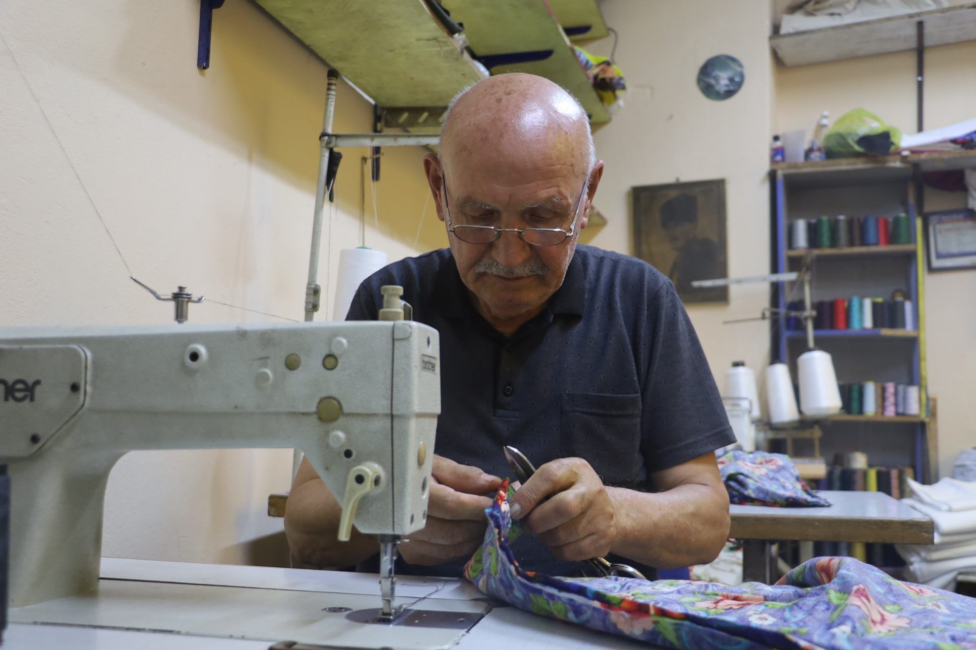 Yaşlı bir adam, dikiş makinesinin başında renkli desenli kumaş üzerinde çalışıyor.