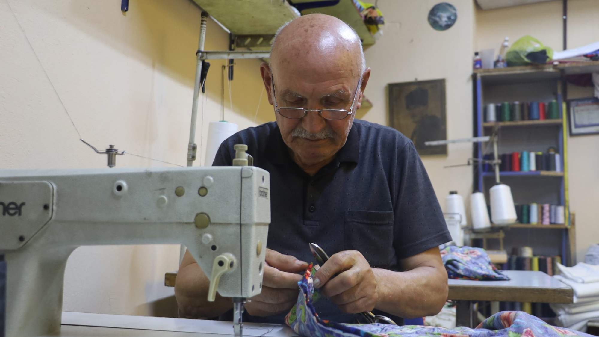 Yaşlı bir adam, dikiş makinesinin başında renkli desenli kumaş üzerinde çalışıyor.