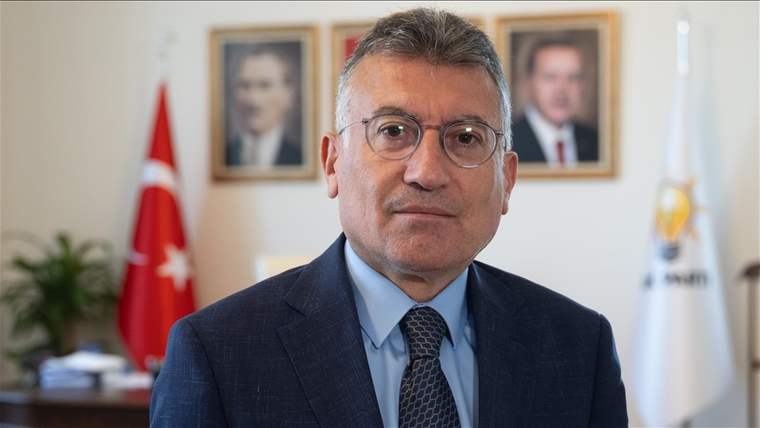 Gözlük takan bir adam, resmi bir kıyafetle poz veriyor. Arka planda Türk bayrağı ve portreler var.