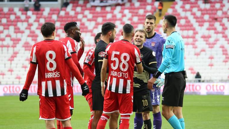 Futbol sahasında kırmızı-beyaz formalı oyuncular, mor formalı rakipleri ve hakemle konuşuyor.