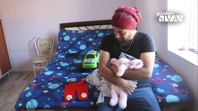 Bir adam, kucağında peluş bir fil tutarak üzgün bir şekilde oturuyor. Yatakta çocuk kıyafetleri ve oyuncak araba var.