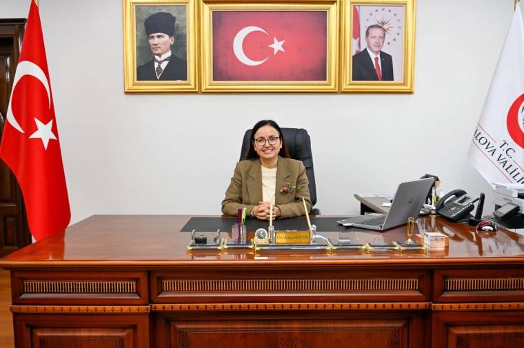 Makam masasında oturan gözlüklü bir kadın, arkasında Türk bayrağı ve çerçeveli portrelerle görülüyor.