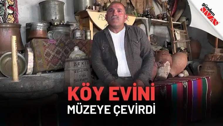 Çeşitli eski ev eşyaları ve kaplarla dolu odada, bir adam sandalyede oturuyor.