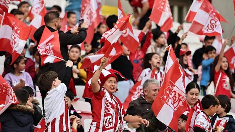 Tribünde kırmızı-beyaz formalı taraftarlar, ellerindeki bayrakları kaldırarak tezahürat yapıyor.