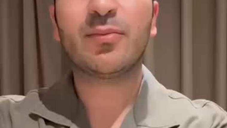 Kısa saçlı bir kişi, açık renkli gömlekle perde önünde kameraya doğru bakıyor.