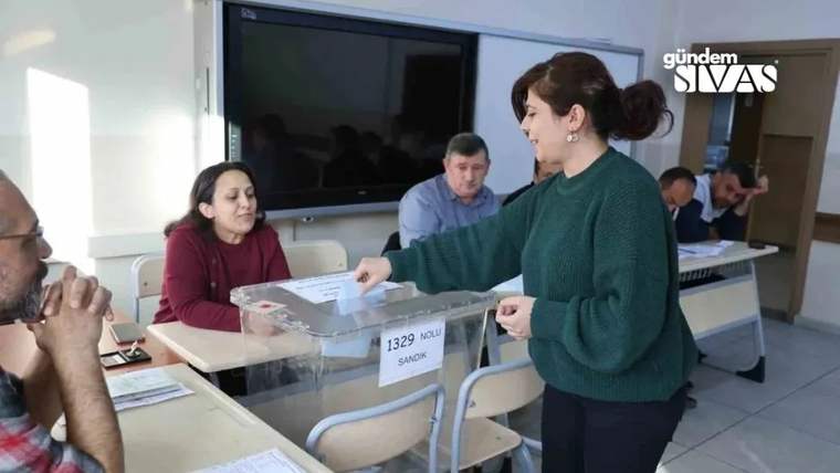 Oy verme işlemi sırasında bir kadın, sandığa oy atıyor; arka planda oturan üç kişi görünüyor.