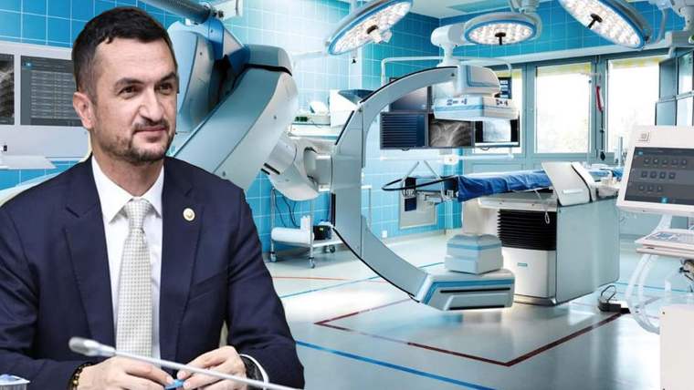 Takım elbiseli bir kişi, ameliyathane ekipmanları ve cerrahi lambaların önünde elinde kalem tutuyor.