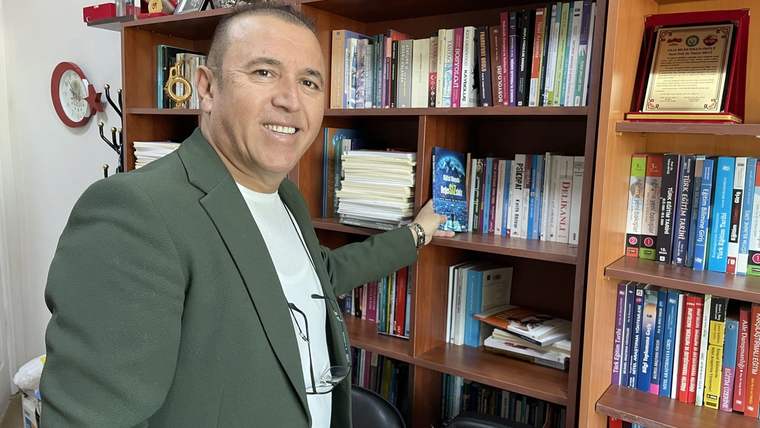 Bir kişi, kitaplık önünde durarak raflardaki kitapları eliyle gösteriyor.