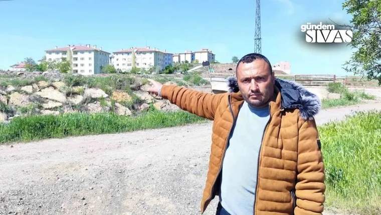 Turuncu montlu bir adam, kayalık arazinin yanındaki toprak yolda sol tarafı gösteriyor; arka planda apartmanlar var.