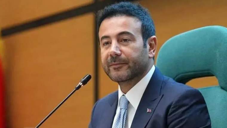 Kürsü önünde oturan takım elbiseli bir erkek, mikrofonun yanında kameraya doğru bakıyor.