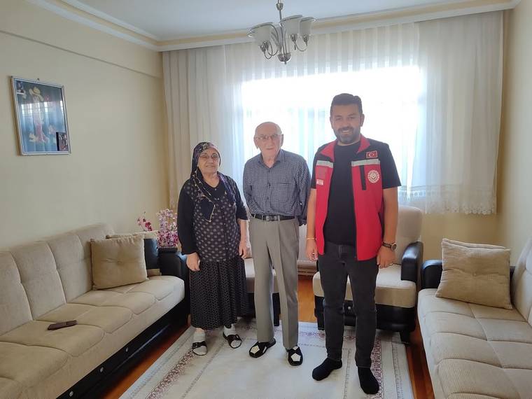 Bir evin salonunda, iki kadın ve bir erkek koltukların önünde yan yana ayakta duruyor.