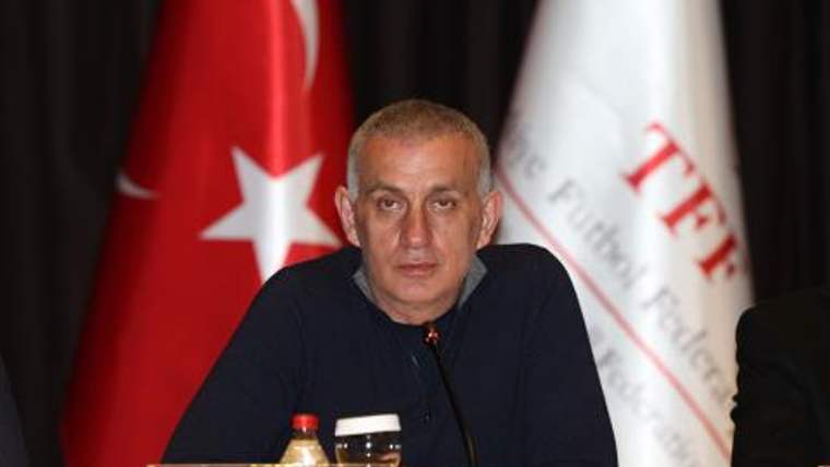 Masa başında mikrofon önünde oturan bir erkek, arkasındaki Türk bayrağı ve başka bir bayrakla görülüyor.