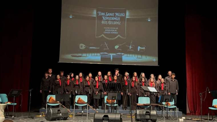 Sahnede siyah kıyafetli bir koro, mikrofon ve müzik aletleri önünde sıralanıyor; arkada konser yazısı bulunuyor.