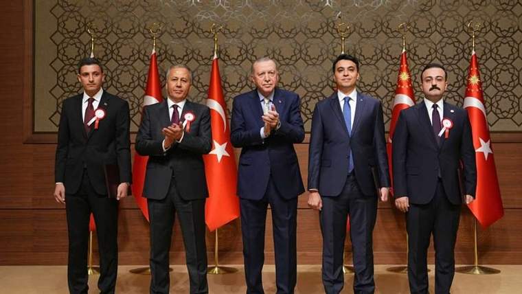 Takım elbiseli beş erkek, arkalarındaki Türk bayrakları önünde duruyor; ortadaki iki kişi alkışlıyor.