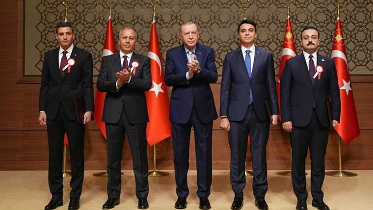 Takım elbiseli beş erkek, arkalarındaki Türk bayrakları önünde duruyor; ortadaki iki kişi alkışlıyor.
