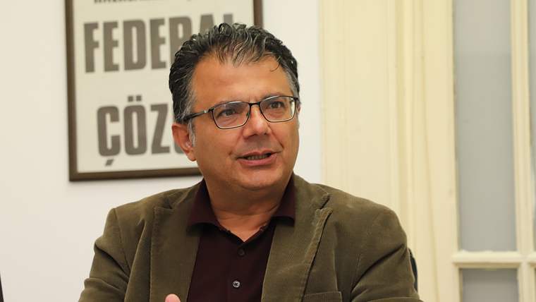 Kahverengi ceket giymiş bir kişi, ofis ortamında oturuyor ve konuşuyor.