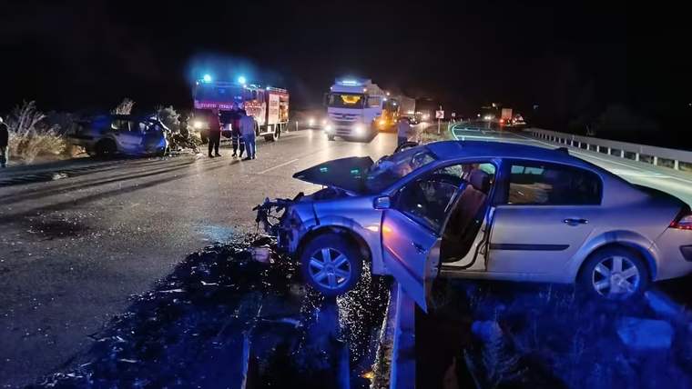 Gece yolunda çarpışmış iki otomobil, açık kapılı gümüş araç ve çevrede duran acil yardım araçları görülüyor.