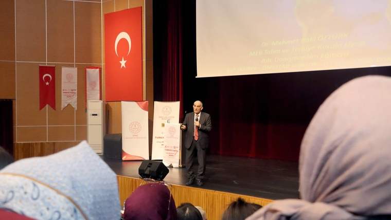Sahne üzerinde konuşan bir kişi, arka planda Türk bayrağı ve afişler var.
