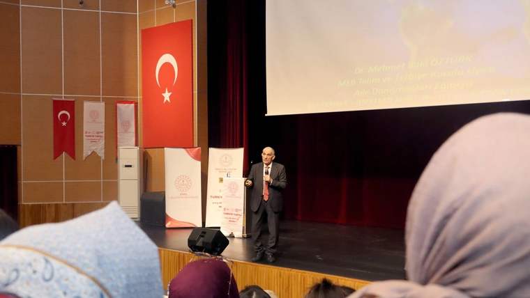 Sahne üzerinde konuşan bir kişi, arka planda Türk bayrağı ve afişler var.