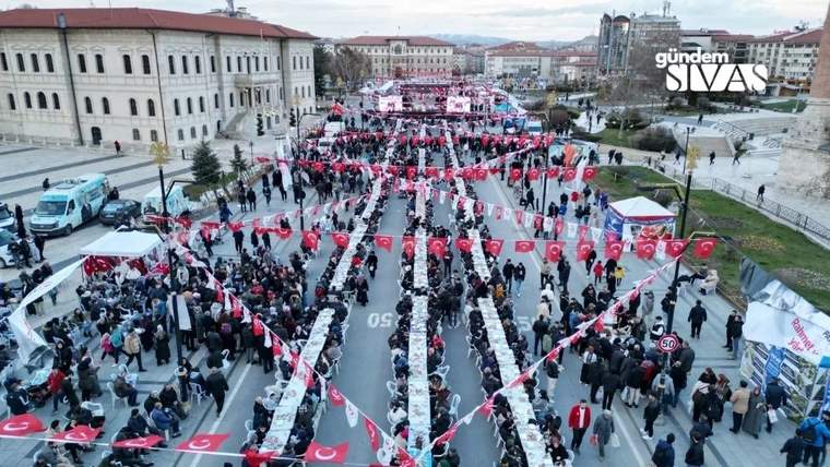 Uzun bir masa düzeninde, Türk bayraklarıyla süslenmiş bir alanda kalabalık bir grup yemek yiyor.