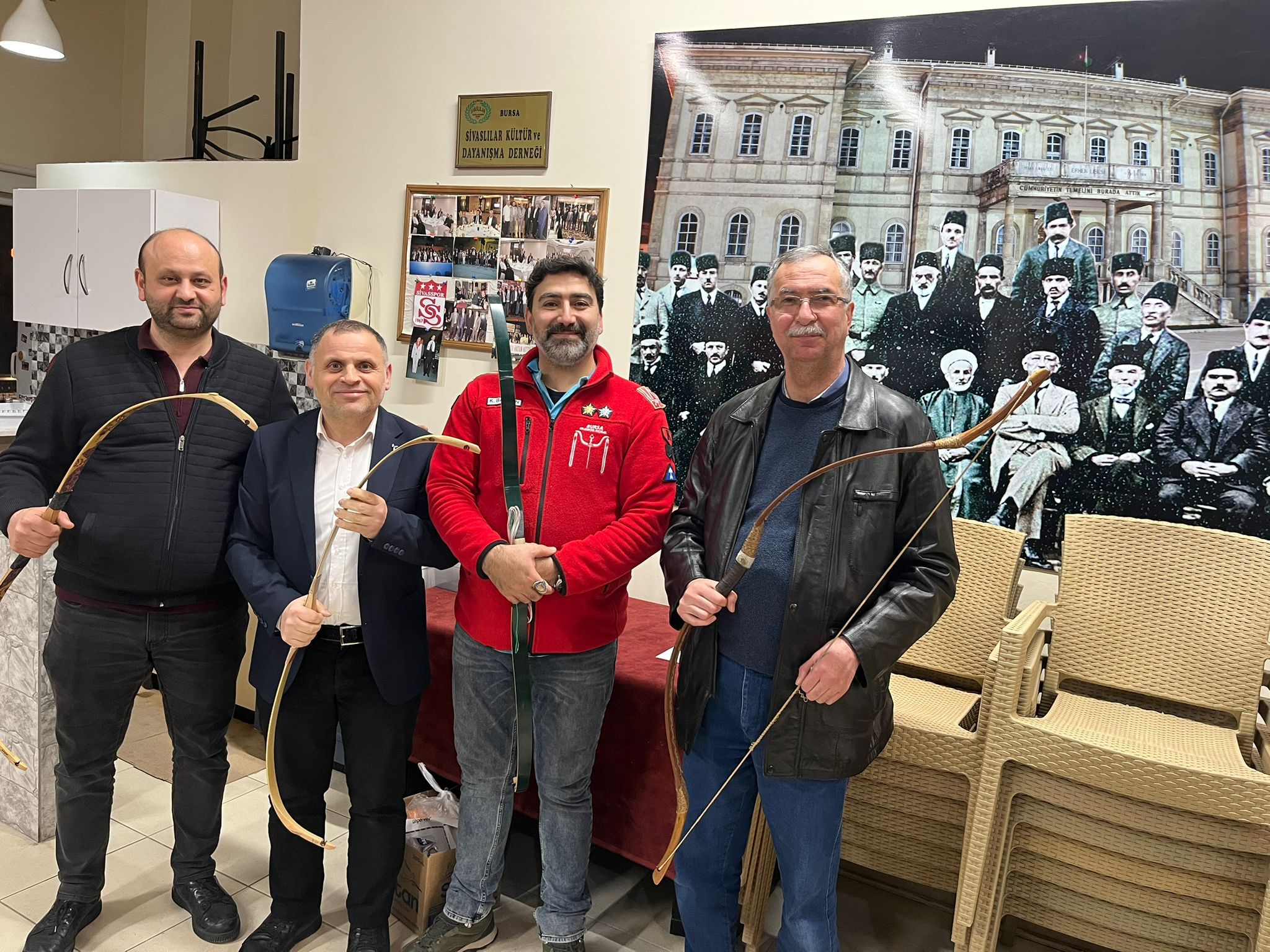 Dört kişi, geleneksel okçuluk malzemeleriyle poz veriyor; arka planda eski bir fotoğraf yer alıyor.