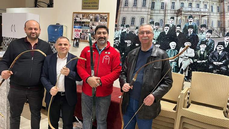 Dört kişi, geleneksel okçuluk malzemeleriyle poz veriyor; arka planda eski bir fotoğraf yer alıyor.