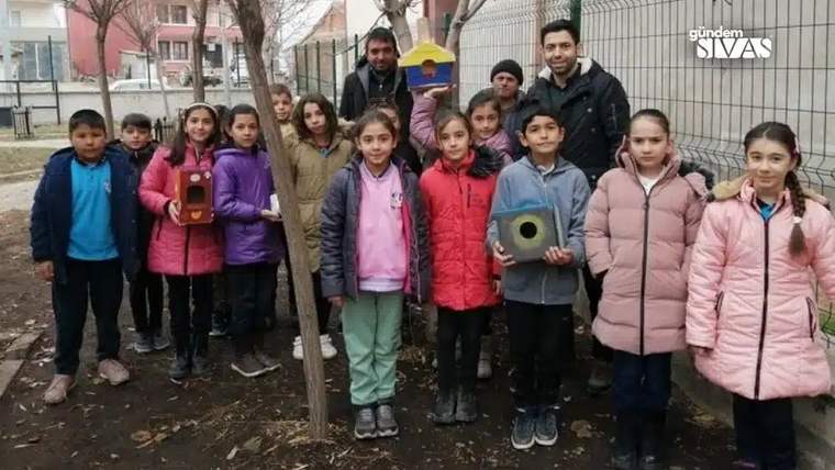 Bir grup çocuk, okul bahçesinde kuş kafesleri ve yemliklerle birlikte ağacın önünde duruyor.