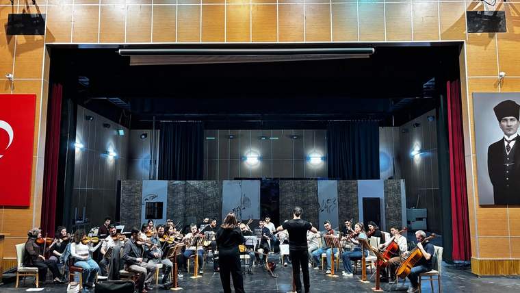 Sahnedeki orkestra üyeleri enstrümanlarıyla otururken, önlerinde iki kişi ayakta duruyor.