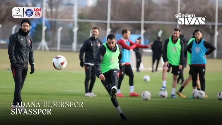 Yeşil yelekli futbolcu antrenman sahasında topa vururken, arka planda diğer futbolcular bekliyor.