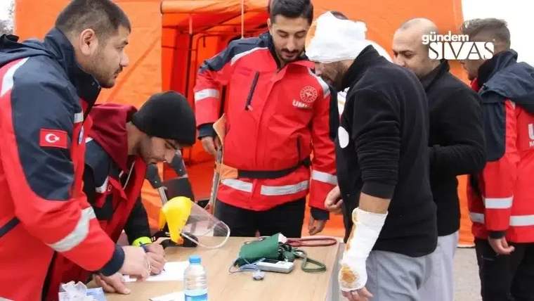 Turuncu çadırda kırmızı üniformalı görevliler, kolu bandajlı bir adamın yanında tıbbi malzemeleri inceliyor.
