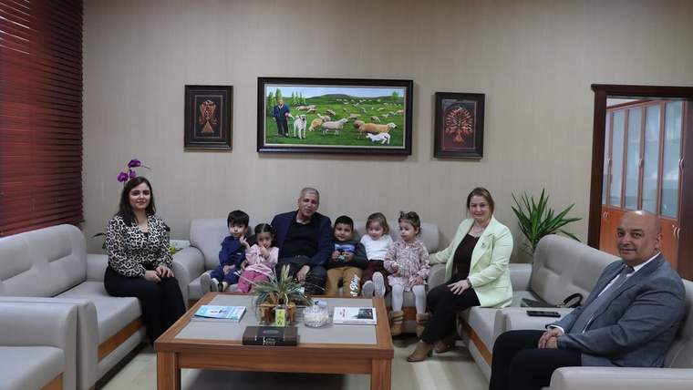 Bir ofiste koltuklarda oturan yetişkinler ve çocuklar, sehpanın önünde birlikte poz veriyor.