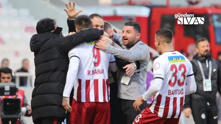 Kırmızı-beyaz formalı futbolcular ve diğer kişiler, saha kenarında birbirine sarılarak sevinç yaşıyor.
